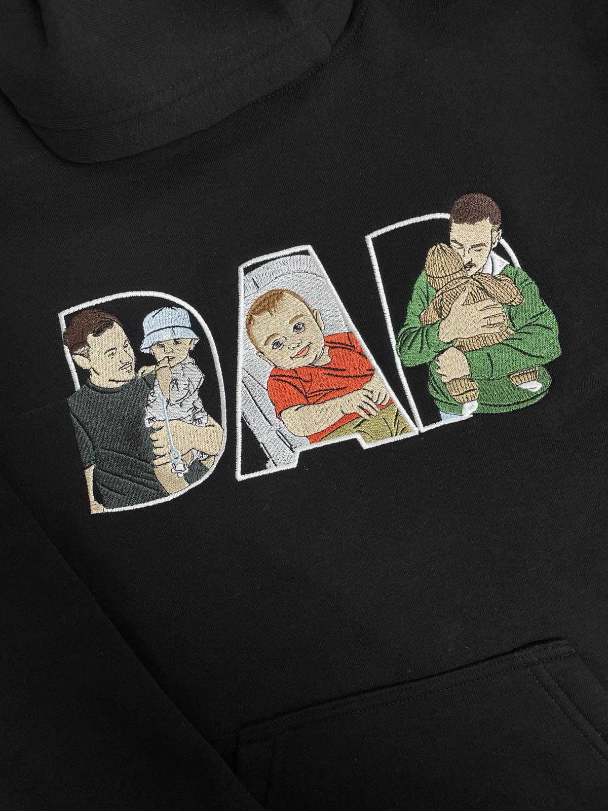 Detaliu de broderie DAD pe hanorac negru, cu un tată și copilul său în fiecare literă